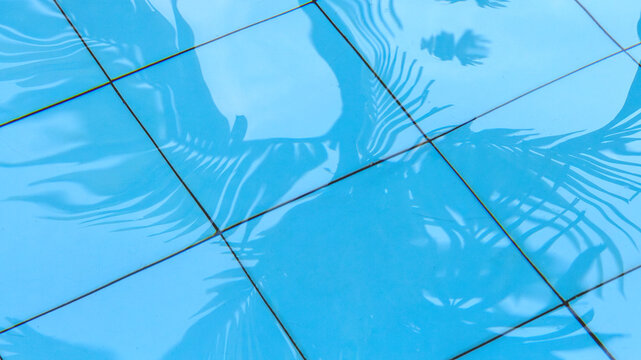 Abstract tropical background featuring palm tree leaf shadows reflecting on clear blue swimming pool tiles through rippling water.