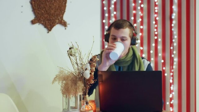 Young caucasian man drinking coffee at laptop, headphones and green scarf, vase with dried flowers, candle and string lights, focused typing and occasional sip, cozy remote workspace for creative