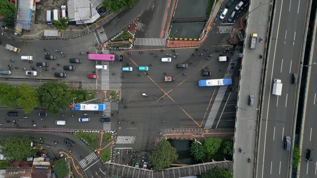 Aerial top-down view of busy X-shaped intersection in Jakarta, Indonesia. Urban traffic with cars, motorbikes, buses, crosswalks, and elevated highway infrastructure.