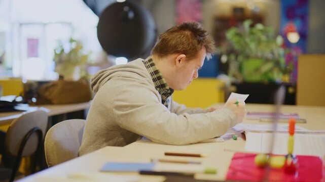 A young student with Down Syndrome diligently reads documents and uses a laptop. He is focused on his studies in a busy learning environment alongside other students.