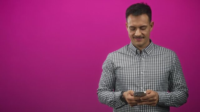 Man with moustache wearing checked shirt holds smartphone with both hands and looks down in studio against magenta backdrop; calm focus.