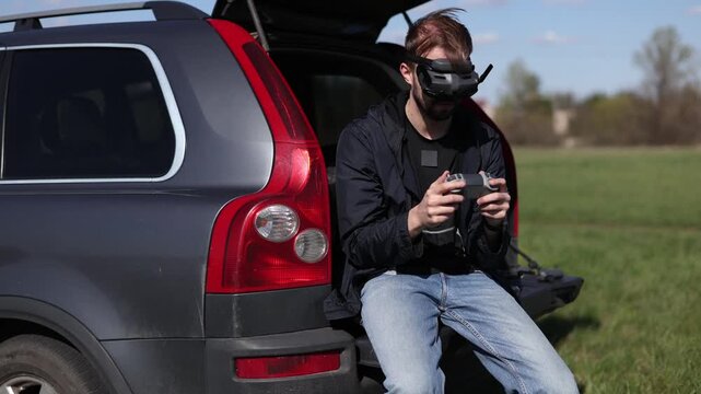 Young man sitting in open SUV trunk in green field wearing FPV goggles and holding remote controller. Person piloting drone using digital headset and transmitter on sunny day in countryside.