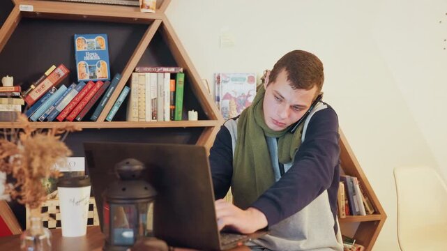 White man on phone at laptop in bright study, wearing green scarf and steady expression, gesturing while speaking, organized bookshelf backdrop, coffee cup and lantern on desk, teleconference vibe,