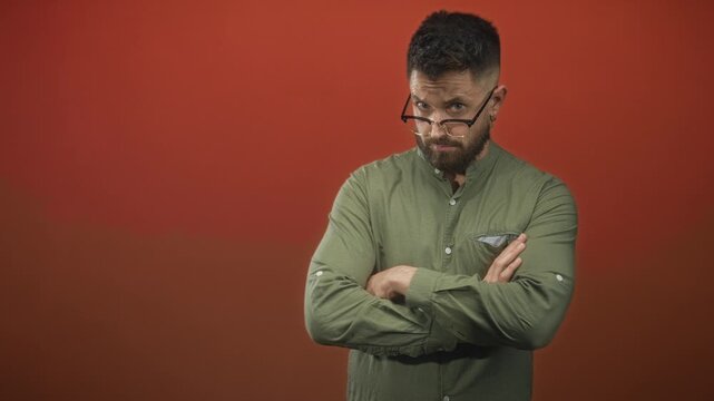 Man adjusts glasses, pushes them down and touches nose with hand, then folds arms while peering over frames in a green shirt before a red studio backdrop; subtle skepticism.