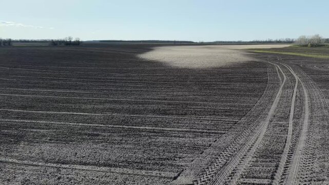 Drone shot showing circular tire patterns on plowed farmland. High contrast between dark soil and large light eroded patches under clear sky. Soil degradation problem.