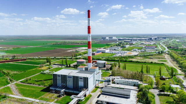 Aerial view of a tall red and white chimney towering over an industrial complex amidst lush green fields under a vast blue sky, Zrenjanin, Vojvodina, Serbia.