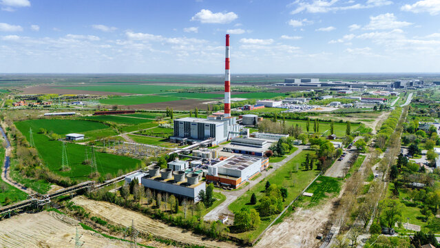 Aerial view of a towering red-and-white striped chimney rising above the industrial complex, contrasting against the patchwork of green fields and a blue sky, Zrenjanin, Vojvodina, Serbia.