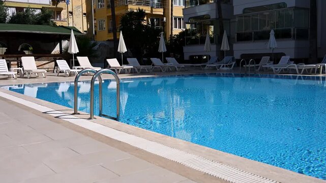 Blue swimming pool at luxury apartment complex or hotel. Sunny resort area with white sunbeds, umbrellas and metal ladder. Summer vacation, hospitality and real estate investment concepts, travel.
