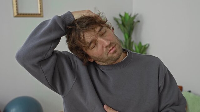 Man performing neck stretch with hand on head and closed eyes beside an exercise ball in a studio; neck mobility recovery relaxation.