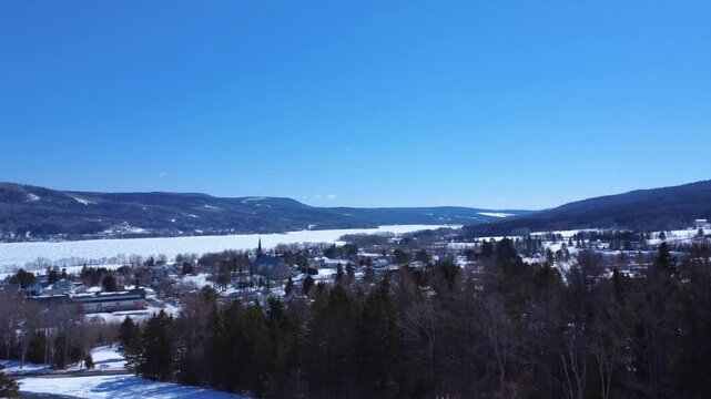 A smooth drone ascent reveals Lac-au-Saumon village and its church, opening to a wide panorama of the Matapedia Valley mountains under a clear blue sky. A serene aerial view of rural Quebec.