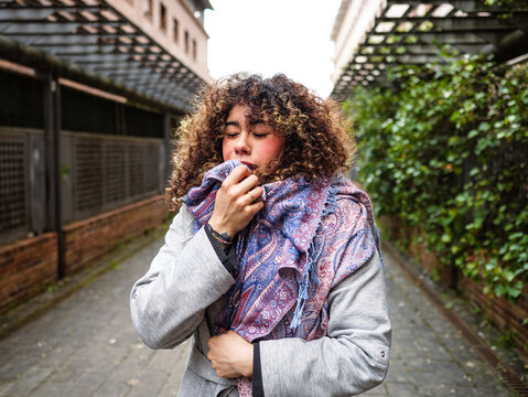 Woman sneeze in cold and flu season, wear coat and scarf in the street. Outdoor portrait of curly hair caucasian girl. Health and illness concept. Allergy symptoms