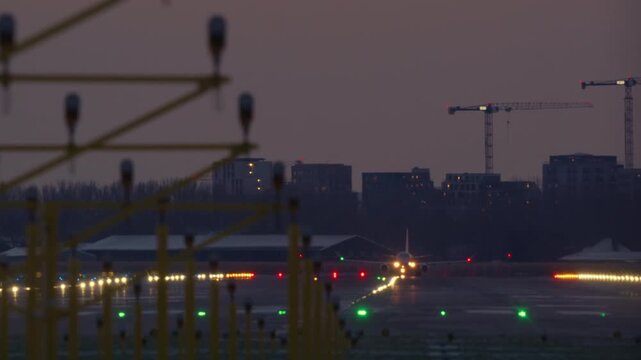 An airplane with bright lights accelerates down a runway and takes off at dusk. The airport is near a city skyline with construction cranes. View from behind the approach lighting system