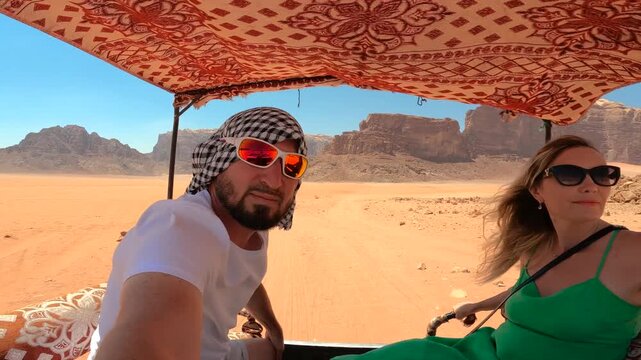 A man in a keffiyeh and a woman in a green dress traveling in the back of a vehicle across the red sand valley of Wadi Rum, Jordan.