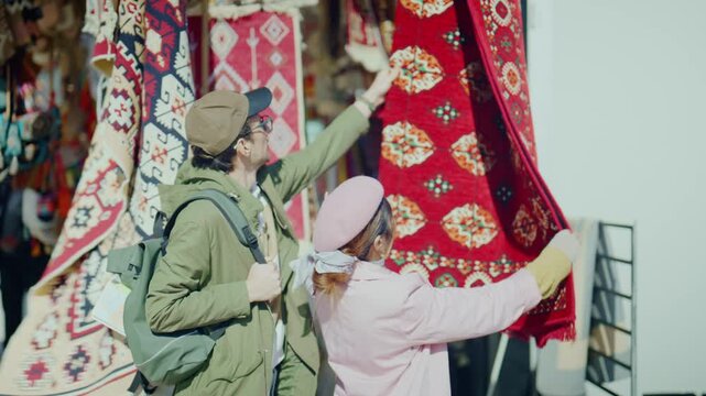 A diverse couple of tourists are seen happily shopping for traditional carpets in an outdoor city market. They explore vibrant textiles, appreciating the cultural crafts and designs.