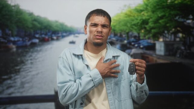 Young hispanic man holding a car keyfob, hand pressed to chest on a street canal bridge; concern uncertainty about a lost or broken car key.