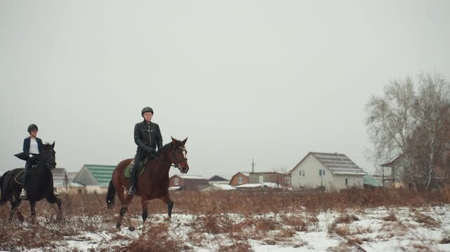Pair of horsemen traverse cold winter landscape. Caucasian pair on horseback across frosty open countryside. Two individuals on horses exploring expansive winter terrain with village skyline