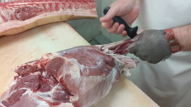 Male butcher in protective gloves skillfully cuts pork meat on a wooden table, with additional meat cuts and a clean kitchen environment visible in the background