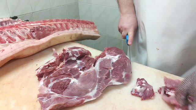 Male butcher in gray apron skillfully cuts pork meat on wooden table, with large pork carcass and neatly arranged meat pieces visible in the background