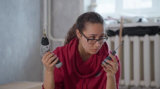 Woman Assembling Furniture Inspecting Electric Drill, Closeup Of Puzzled Hands Holding Drill And Screwdriver, Indoor Apartment Renovation Mood, Soft Daylight, Radiator And Tools In Background, Focused