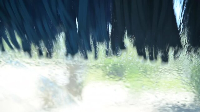 Point of view video from inside a car during an automatic car wash. Brushes rotate across the windshield while water, foam and droplets move and distort the view, creating a dynamic immersive scene