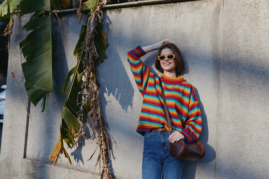 Woman wearing a colorful striped sweater and sunglasses leaning against a concrete wall with tropical leaves, smiling in a casual outfit and jeans in an urban lifestyle travel scene.