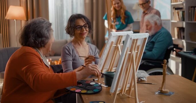 Two senior women painting together on canvas in assisted living home, smiling and talking. Warm moment of friendship, creativity and social interaction in relaxed indoor setting.