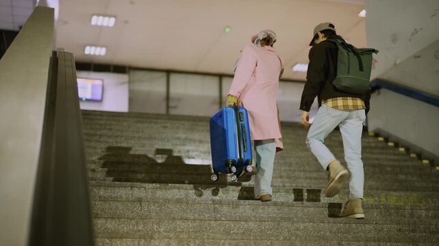 A young couple climbs concrete stairs, the woman pulling a suitcase while the man wears a backpack. Two travelers ascend public steps with their belongings, navigating an urban environment.