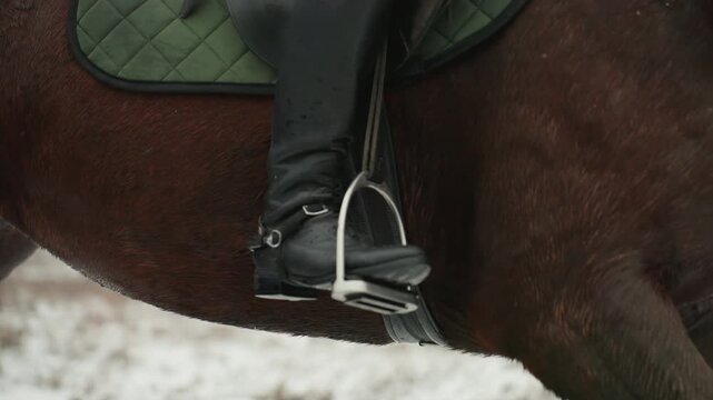 Saddle closeup rider leg in snow, mounted training moment focusing on stirrup, girth and leather tack, rider balancing posture, green pad visible, snowy rural background, instructional tone