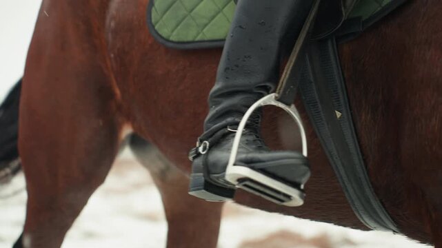 Closeup stirrup boot on horse pad gleaming steel stirrup cradles black leather boot with green saddle pad visible, snowflakes dust coat, focus on grip balance and strap tension, equestrian equipment