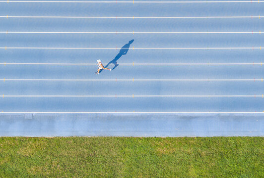 Aerial view of a lone runner jogging on a blue athletic track with white lane lines and a long shadow cast on the surface next to a strip of green grass in Taiwan.