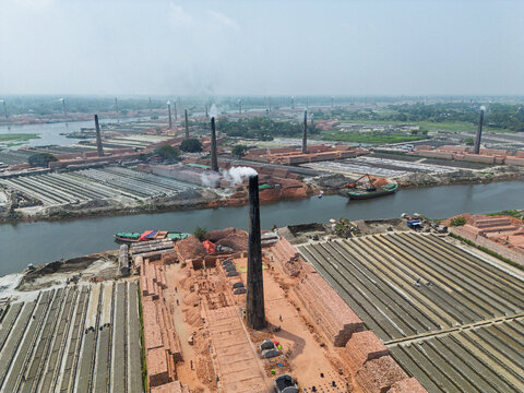 Aerial view of traditional brick kilns with tall chimneys emitting smoke and rows of drying bricks along the riverbank in Narayanganj, Dhaka Division, Bangladesh.