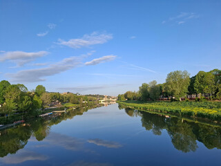 Obraz premium Peaceful river view with green trees, clear blue sky and soft cloud reflection on still water. Natural scenic panorama ideal for travel, relaxation and summer themes.