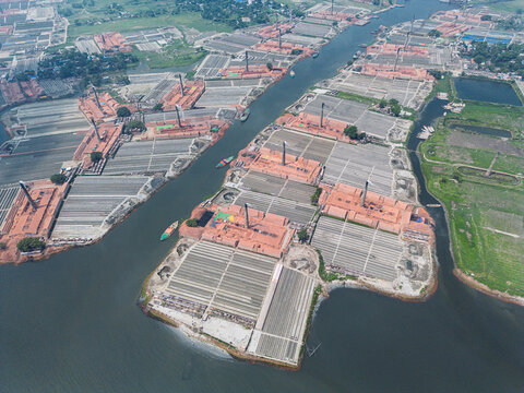 Aerial view of traditional brick kilns with tall chimneys and rows of drying bricks along the riverbank in Narayanganj, Dhaka Division, Bangladesh.