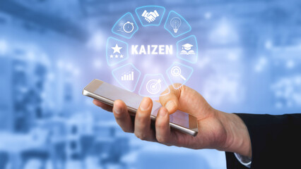 Fototapeta premium Hand Holding Smartphone Displaying Kaizen Concept with Growth Icons and Blue Background in a Modern Office Setting for Business Strategy and Improvement Quark.
