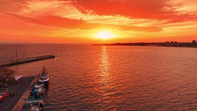Setting sun colors the sky and waterscape with bright orange light. Drone footage rising over the pier with boats standing by. Nessebar, Bulgaria.