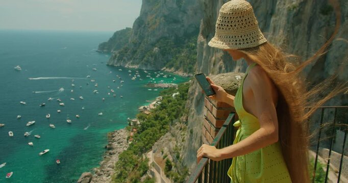 A woman views Marina Piccola from a cliffside lookout while using her phone on a summer day in Capri Italy