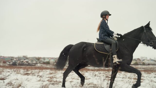 Woman guides horse through snow. Horse and rider pass snowy landscape during training session. Female equestrian on horseback practicing trot along city outskirts in winter conditions