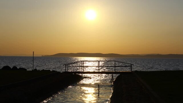 sunset light reflecting on moving lake balaton surface with soft wave patterns and a small foreground bridge creating a layered view of water, glow and shoreline silhouettes