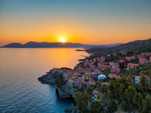 Aerial view of the colorful houses of Tellaro perched on rocky cliffs during a vibrant sunset over the calm sea in Tellaro, Liguria, Italy.