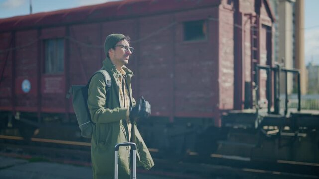 A young male tourist stands patiently on an urban train station platform with his luggage. He checks his watch, contemplating his journey amidst old train wagons.
