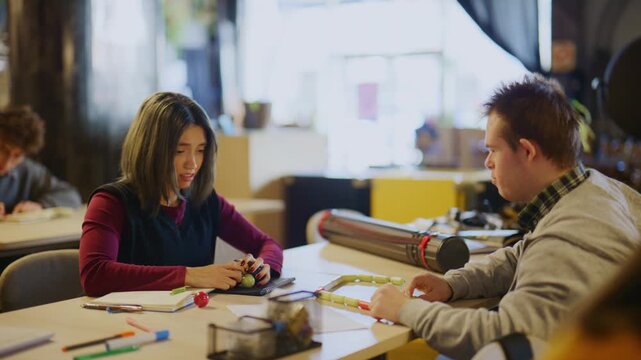 Students, including one with Down syndrome, collaborate on a learning activity at a desk. Other students are seen studying and focusing on their tasks in the background.