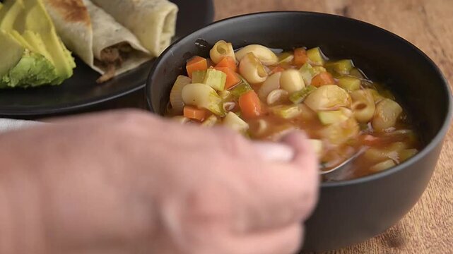 Elbow or macaroni pasta soup with tomato and chopped vegetables seasoned with chicken broth and spices, topped with goat cheese and chipotle chili, in a black bowl, with a plate of machaca burritos 