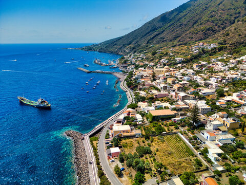 Aerial view of Salina island coastal town with a harbor, anchored ship, and lush green hills under a clear blue sky in Metropolitan City of Messina, Sicily, Italy.