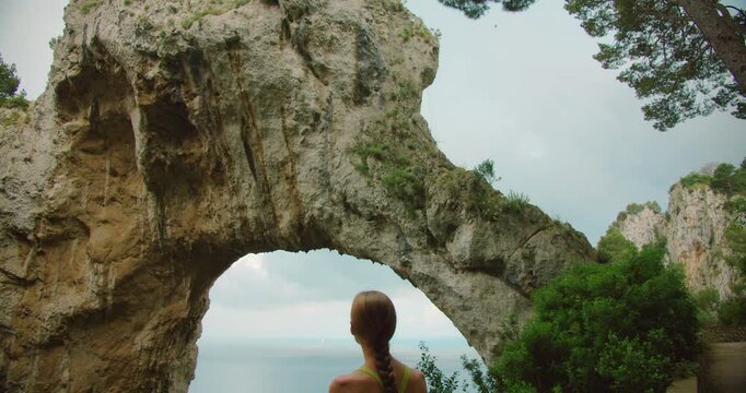 A woman holding a map admires the Arco Naturale and sea view while hiking a summer trail in Capri Italy