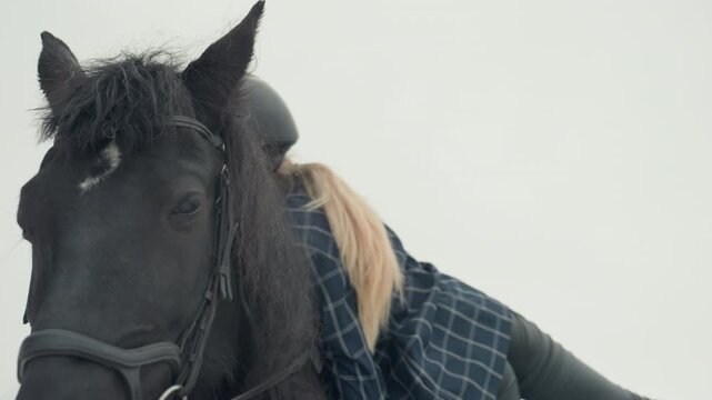 Helmeted woman tending black horse in snowy paddock during handson tandem training, adjusting bridle and stroking mane, close interaction, calm expression, outdoor stable setting