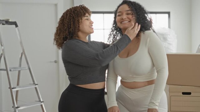 Woman smiles, hugs and kisses her friend's cheek beside stacked moving boxes in a sunlit apartment; friendship joy celebration.