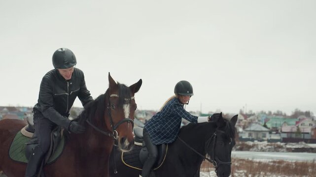 Two caucasian riders practicing near village, tandem education exercise with partners working rhythm and balance, canter and trot over snowy field, houses on horizon and focused teamwork