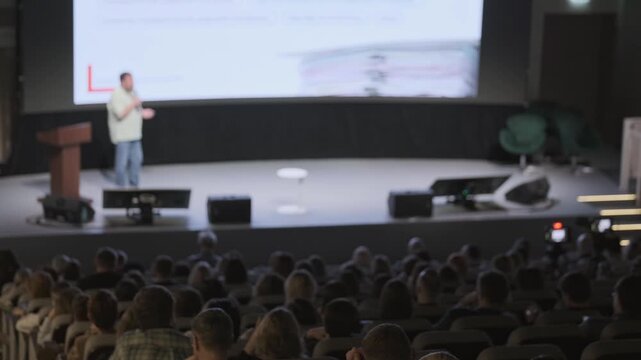 Speaker presenting on stage to large audience at tech conference, blurred foreground shows attendees watching presentation and slides on projection screen.