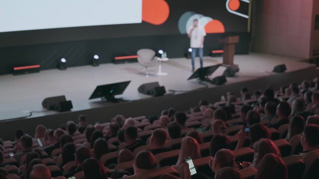 Crowded auditorium with audience listening to speaker on stage during tech conference presentation, dim lighting and mobile phones visible.