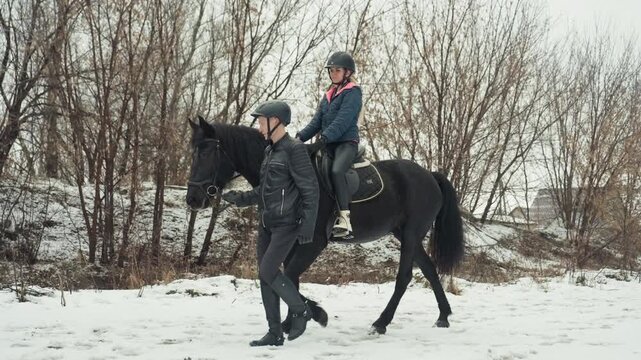 Instructor leading helmeted rider on horse, close framing shows hand guiding reins and steady pace, patient mentor walking beside dark mount, snowy thicket and village roofs in background, safety gear